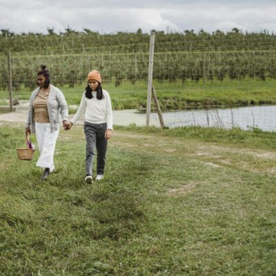 Ethnic mother with wicker basket holding hand of daughter wearing while walking from fence separating lake and plantation