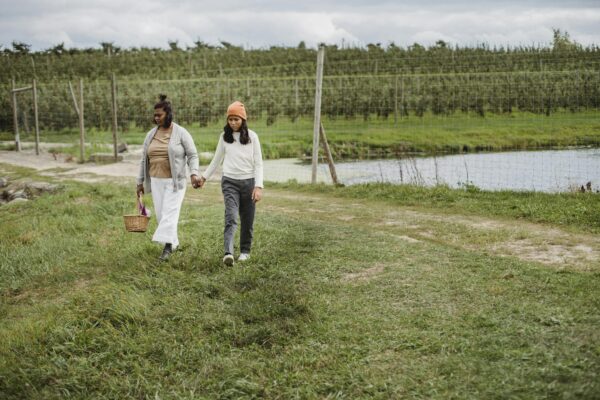 Ethnic mother with wicker basket holding hand of daughter wearing while walking from fence separating lake and plantation