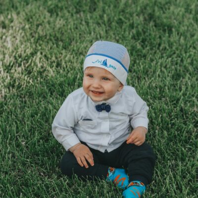 Full body of adorable child in formal wear and hat smiling and sitting on green grassy meadow at daylight