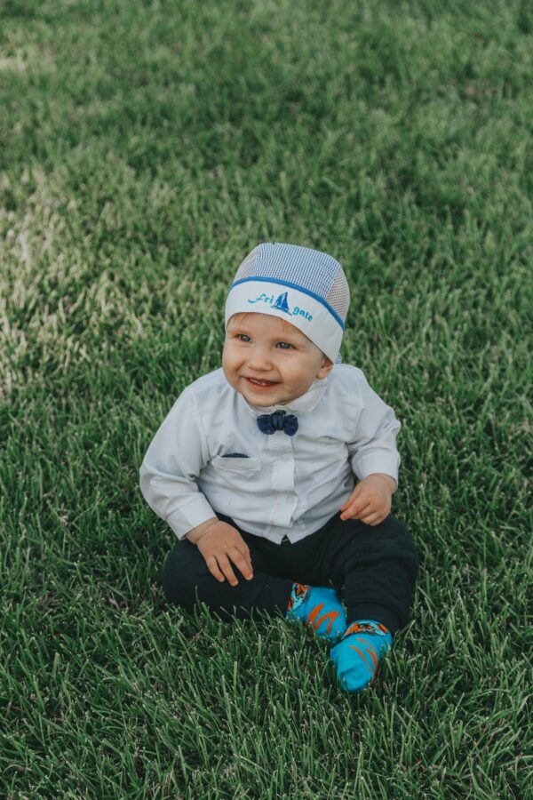 Full body of adorable child in formal wear and hat smiling and sitting on green grassy meadow at daylight